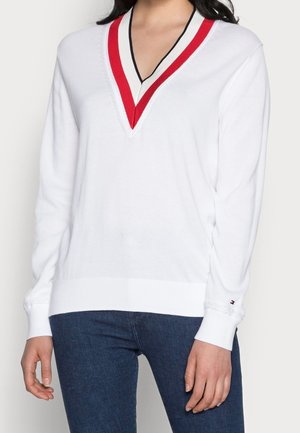 Femme portant un pull blanc à col en V avec des rayures rouges et noires au niveau du col et un jean bleu, vue des épaules jusqu'aux hanches.