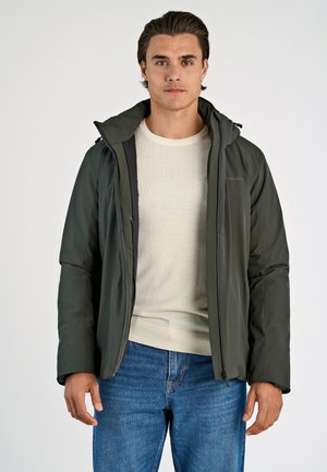 Dunkelgrüne isolierte Jacke mit Kapuze, Reißverschluss und elastischen Bündchen, getragen über einem cremefarbenen Pullover und Jeans. Glatte Textur, minimale Details.