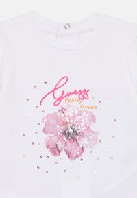 Λευκό βαμβακερό μπλουζάκι με κοντά μανίκια, με σχέδιο ροζ παγιέτας σε σχήμα λουλουδιού και κείμενο "Guess Pretty Flower" σε ροζ και πορτοκαλί.