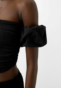 Haut court noir présentant un design texturé froncé et des manches courtes bouffantes. Le tissu est léger avec une finition lisse.