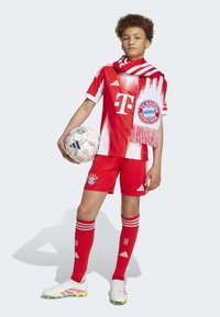 Mládežnícky futbalový dres Bayernu Mníchov: červený dres s bielymi akcentmi, šortky, červené ponožky s bielymi prúžkami, biele kopačky, drží futbalovú loptu.