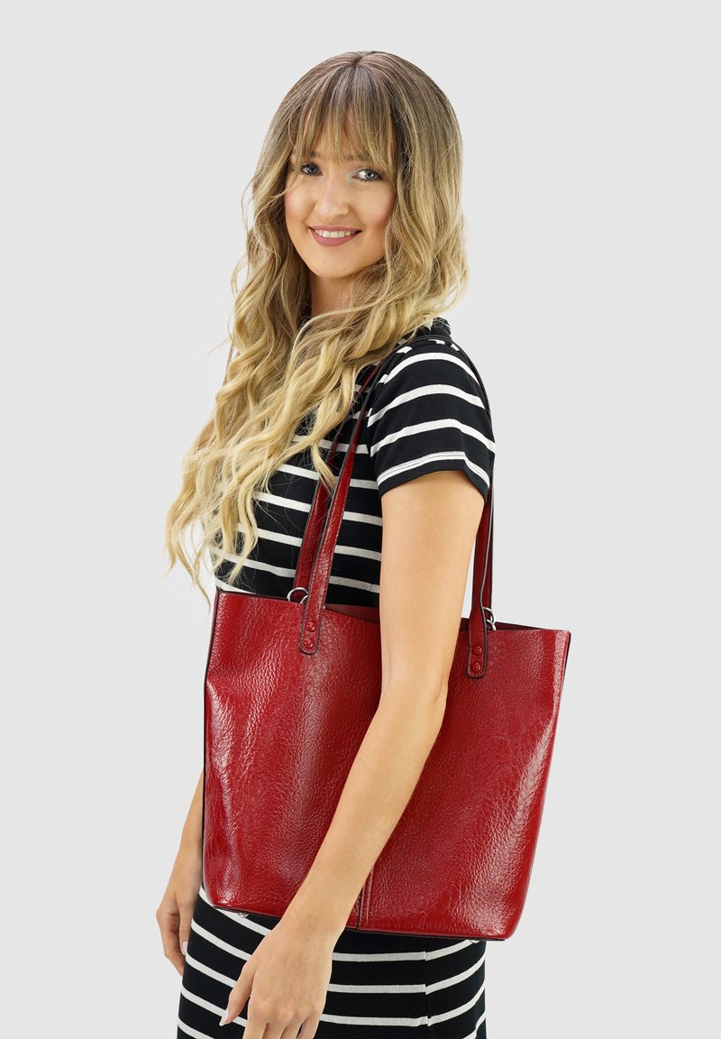 Vrouw met lang golvend blond haar, gekleed in een zwart-wit gestreepte jurk, met een grote glanzende rode leren schoudertas.