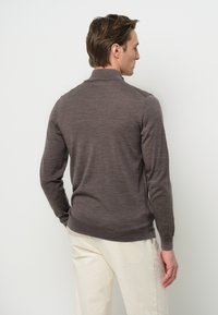 Brauner Strickpullover mit hohem Kragen, gerippten Bündchen und Saum. Kombiniert mit hellfarbigen Hosen. Weiche Textur, minimalistisches Design.