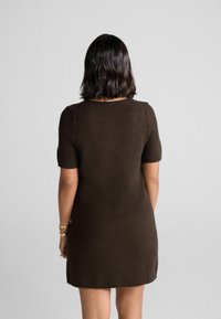 Frau mit schulterlangem, dunklem Haar, die ein kurzärmliges, dunkelbraunes, knielanges Kleid trägt und vor einem schlichten, hellen Hintergrund steht.