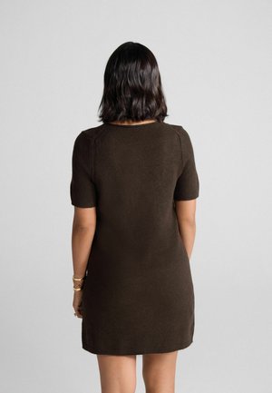 Frau mit schulterlangem, dunklem Haar, die ein kurzärmliges, dunkelbraunes, knielanges Kleid trägt und vor einem schlichten, hellen Hintergrund steht.