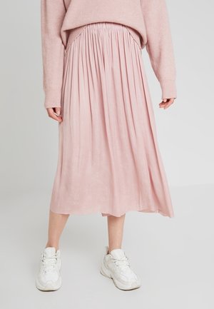 Femme portant une jupe midi plissée rose, un pull rose assorti et des baskets blanches épaisses, debout devant un fond uni.