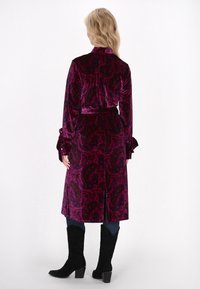Samtmantel in tiefem Violett mit Paisleymuster, hohem Kragen und Schleifenakzenten an den Ärmeln; kombiniert mit schwarzen kniehohen Stiefeln und Jeans.