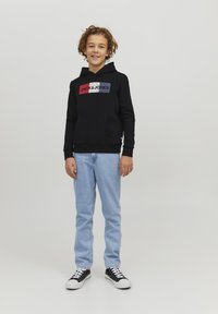Felpa nera con un logo centrale multicolore, abbinata a jeans blu chiaro e sneakers nere con suola bianca, in piedi di fronte a uno sfondo neutro.