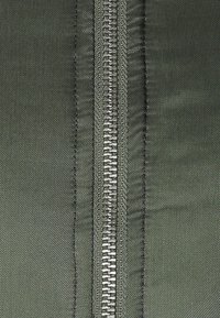 Grön dragkedja på tyg, med en metallisk yta och tätt placerade tänder. Slät, texturerad omgivande textil med en subtil lyster.