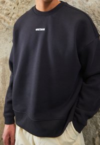 Personne portant un sweat-shirt noir avec le texte blanc "WRSTBHVR" sur la poitrine, debout contre un mur en béton texturé, la main dans la poche.