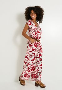 Jumpsuit floral rojo y blanco con mangas cortas, escote en V, piernas anchas y un dobladillo con estampado, combinado con sandalias de tacón marrones.
