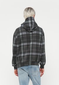 Sudadera con capucha de cuadros en negro y gris, de corte holgado y con puños acanalados. Las características notables incluyen una capucha oscura y líneas de color sutil en el patrón.