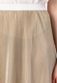 Pleated skirt in a shimmering beige fabric with a textured, ribbed design and a white elastic waistband. Close-up view highlighting the details.