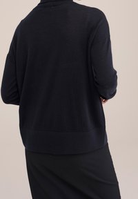 Person som bär en löst sittande svart långärmad tröja och svarta byxor, visad bakifrån mot en enkel beige bakgrund.