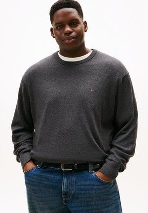 Homme portant un pull gris foncé sur une chemise blanche et un jean bleu, debout les mains dans les poches devant un fond uni.