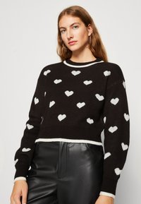 Črni pleteni pulover z belim vzorcem srca, žametnimi manšetami in robom. Okrogel ovratnik z belim pasom. Nošen z črnimi usnjenimi hlačami.