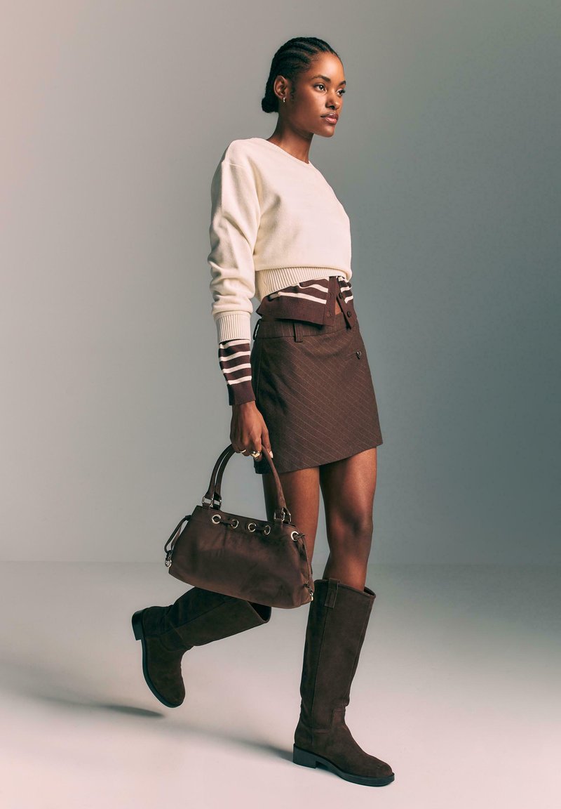 Des bottes en suède marron hautes jusqu'au genou, une mini-jupe à rayures marron, et un pull crème avec des détails à rayures aux poignets. Un sac à main marron assorti est porté.