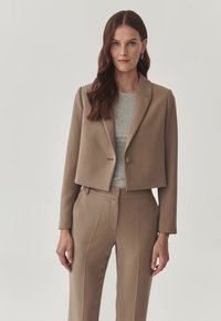 Cropped beige blazer with notch lapels, a single button closure, and smooth texture, paired with gray top, featuring tailored pants.