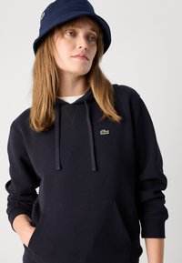 Mujer con un sombrero de cubo azul marino y una sudadera a juego con un pequeño logo de cocodrilo verde, con las manos en el bolsillo de la sudadera, mirando ligeramente hacia la derecha.