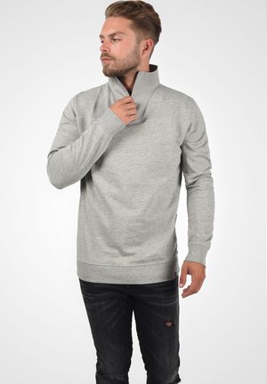 Hellgrauer Hoodie mit hohem Kragen und Reißverschluss, langärmelig und in lässiger Passform. Dunkle Jeans mit Distressed-Details.