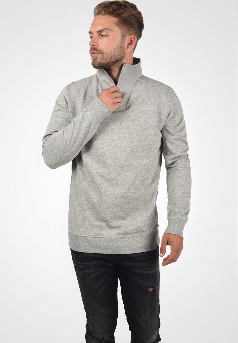 Lys grå hettegenser med høy krage og glidelås, med lange ermer og avslappet passform. Mørke jeans med slitte detaljer.
