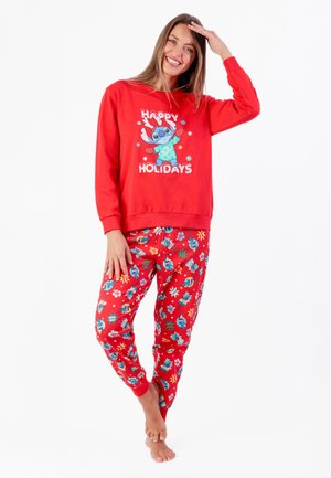 Mujer sonriente con pijama roja temática navideña con dibujo de personaje de dibujos animados y texto "Felices Fiestas", descalza, de pie contra fondo blanco.
