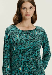Mujer con cabello largo castaño que lleva una blusa de manga larga con un patrón de paisley en color teal, de pie contra un fondo claro y liso.