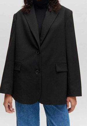 Persona con blazer negro de doble botonadura sobre un suéter de cuello alto negro y jeans azules, de pie frente a un fondo claro y liso.