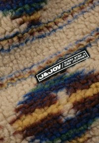 Fleece stof met een beige basis met kleurrijke abstracte patronen in blauw, groen, bruin en geel. J&JOY-label bevestigd.