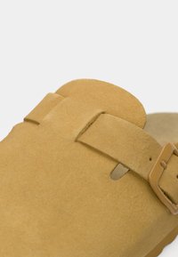 Καφέ σανδάλι από σουέτ με ρυθμιζόμενο λουράκι και μεταλλικό πόρπη. Διαθέτει απαλή υφή και ανοιχτή μύτη, προσφέροντας άνεση.