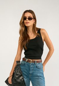 Junge Frau in schwarzem Tanktop, blauen Jeans, braunem Gürtel, Sonnenbrille tragend, hält eine schwarze Ledertasche, posiert vor einfachem Hintergrund.