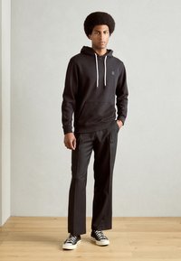 Haut à capuche en coton noir avec cordon de serrage et poche avant, associé à un pantalon noir ajusté et des baskets noir et blanc. Fond uni.