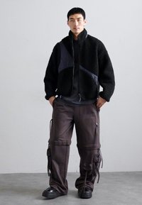 Giacca in pile nera con zip, caratterizzata da un pannello scuro a contrasto. Pantaloni cargo larghi marroni con zip e stringhe, abbinati a scarpe nere.