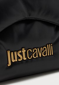 Bolso de mano de cuero negro con un diseño texturizado y acolchado y el logo dorado de "Just Cavalli" exhibido de forma prominente. Textura suave, estilo minimalista.