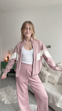 Světle růžový teplákový komplet s bílými diagonálními pruhy na bundě a pase; má volný střih a měkkou látku. Model nosí bílé tílko.