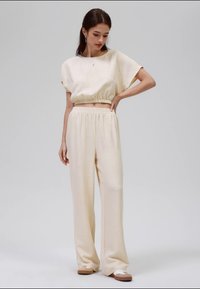Mulher de pé com a mão na cintura, vestindo calças largas texturizadas de cor creme e uma blusa curta de manga curta, acompanhadas de ténis brancos.