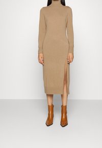 Beiges Strickkleid mit langen Ärmeln und hohem Ausschnitt, mit einem Seitenschlitz. Kombiniert mit braunen Stiefeletten. Glatte Textur.