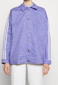 Fialová bunda s bílými vlnkovými vzory, zapínáním na knoflíky vpředu a špičatým límcem. Na každém rukávu vedou tři bílé pruhy.