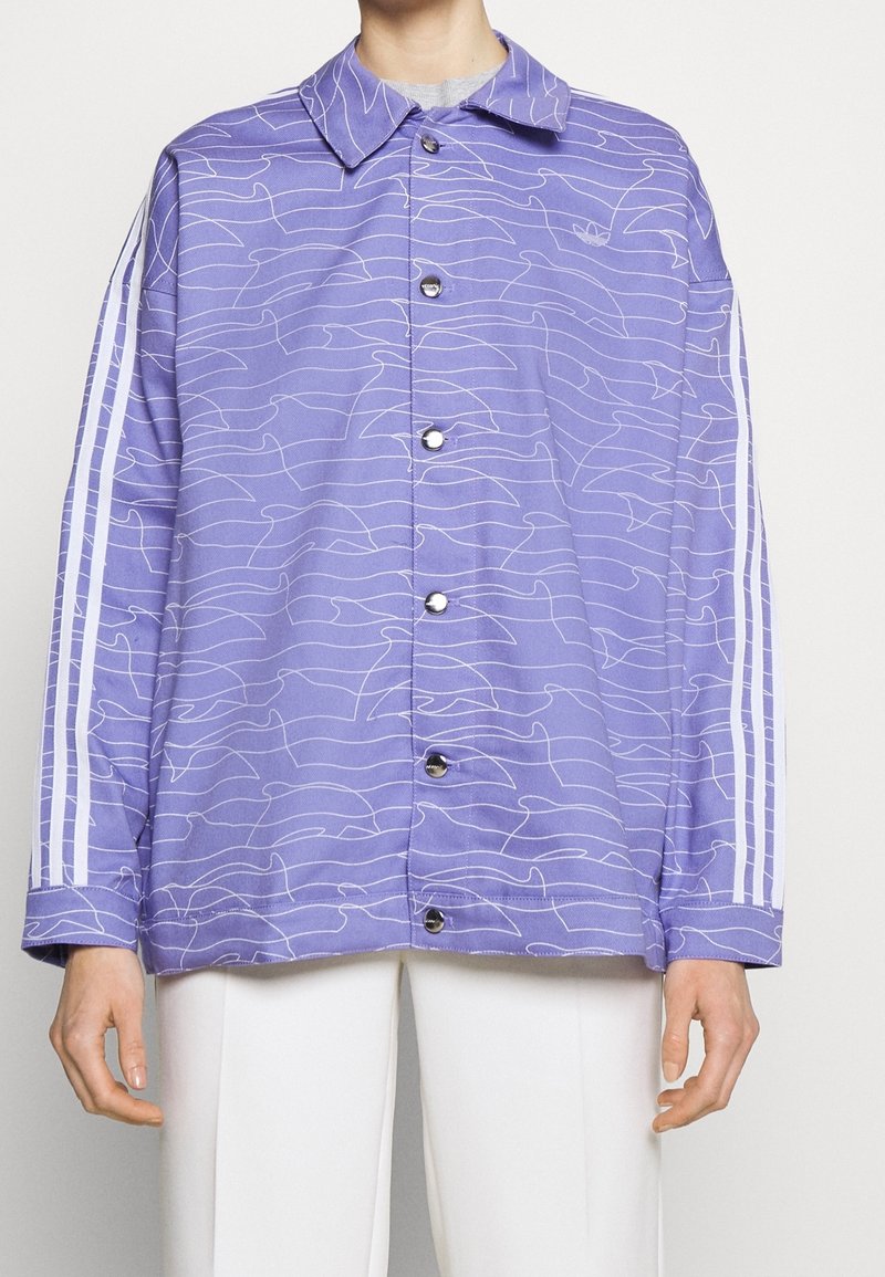 Fialová bunda s bílými vlnkovými vzory, zapínáním na knoflíky vpředu a špičatým límcem. Na každém rukávu vedou tři bílé pruhy.