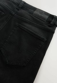 Melnas d�žinsu bikses ar taisnu griezumu, divām aizmugures kabatām un faktūras ādas uzšuvumu uz jostasvietas ar iespiestu tekstu.