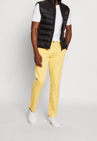 Un uomo si erge con sicurezza, indossando un gilet imbottito nero sopra una camicia bianca, abbinato a pantaloni gialli e sneakers bianche, su uno sfondo neutro.