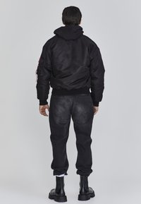 Man die met zijn rug naar de camera staat, gekleed in een zwarte leren bomberjack met capuchon, vervaagde zwarte jeans, witte sokken en zwarte leren laarzen, op een grijze achtergrond.