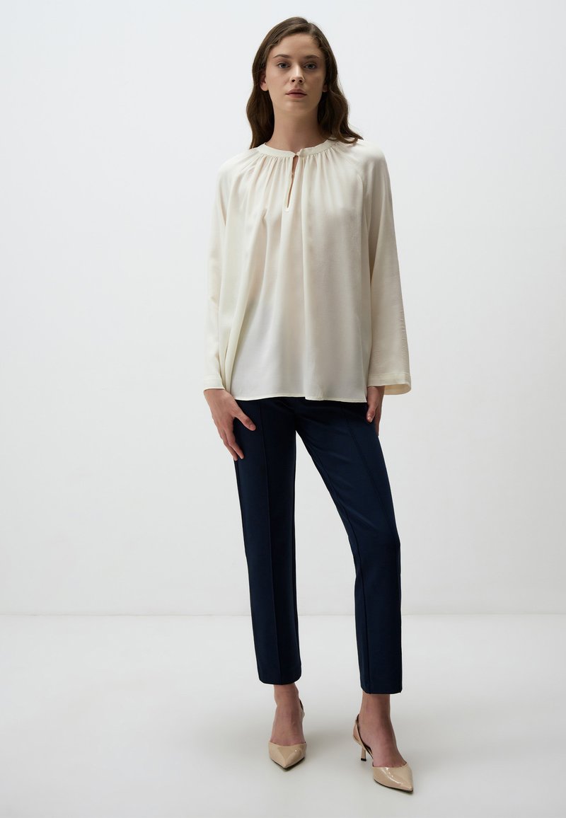 Blouse crème avec un col froncé et des manches longues, associée à un pantalon tailleur bleu marine et des escarpins beiges à bout pointu. Texture de tissu lisse.