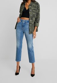 Camo-print green jacket, black cropped top, and high-waisted blue jeans with a straight leg. Black high-heeled shoes accentuate the outfit.