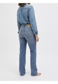 Ljusblå denimjeans med rak bendesign, fem fickor och en läderlapp vid midjan. En enkel blå skjorta med uppvikta ärmar.