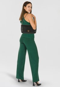 Schwarze Lederhandtasche mit strukturierter Oberfläche und goldfarbenen Beschlägen, diagonal getragen. Modell in grünem Outfit, zeigt locker sitzende Hose und Schuhe mit Absatz.