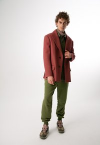 Rdzawy wełniany żakiet z wyciętymi klapami noszony na zielonej koszuli z guzikami. Oliwkowe dresy i wielokolorowe sneakersy dopełniają strój.