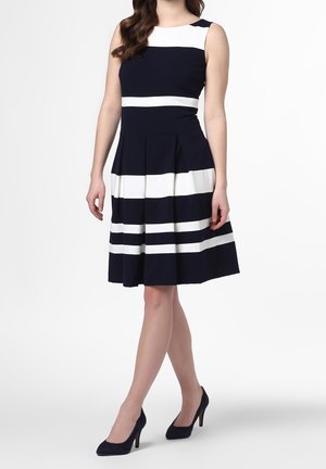 Un vestido azul marino con rayas horizontales blancas, diseño sin mangas, corsé ajustado y falda plegada. Llevado con zapatos de tacón alto azul marino.