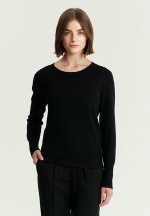 Donna con i capelli castani corti, che indossa una maglietta nera aderente a maniche lunghe e pantaloni neri, in piedi davanti a uno sfondo chiaro e uniforme.