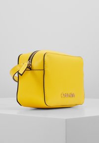 Borsa quadrata in pelle gialla con chiusura a zip, bordi neri e dettaglio logo frontale. Dotata di manico corto per un facile trasporto.